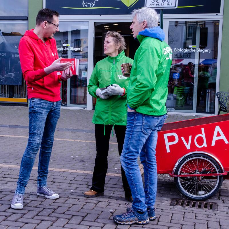 Foto van twee GroenLinks campagnevoerders en een PvdA campagne voerder in het centrum van Hilversum, staand voor een bakfiets met PvdA logo.  